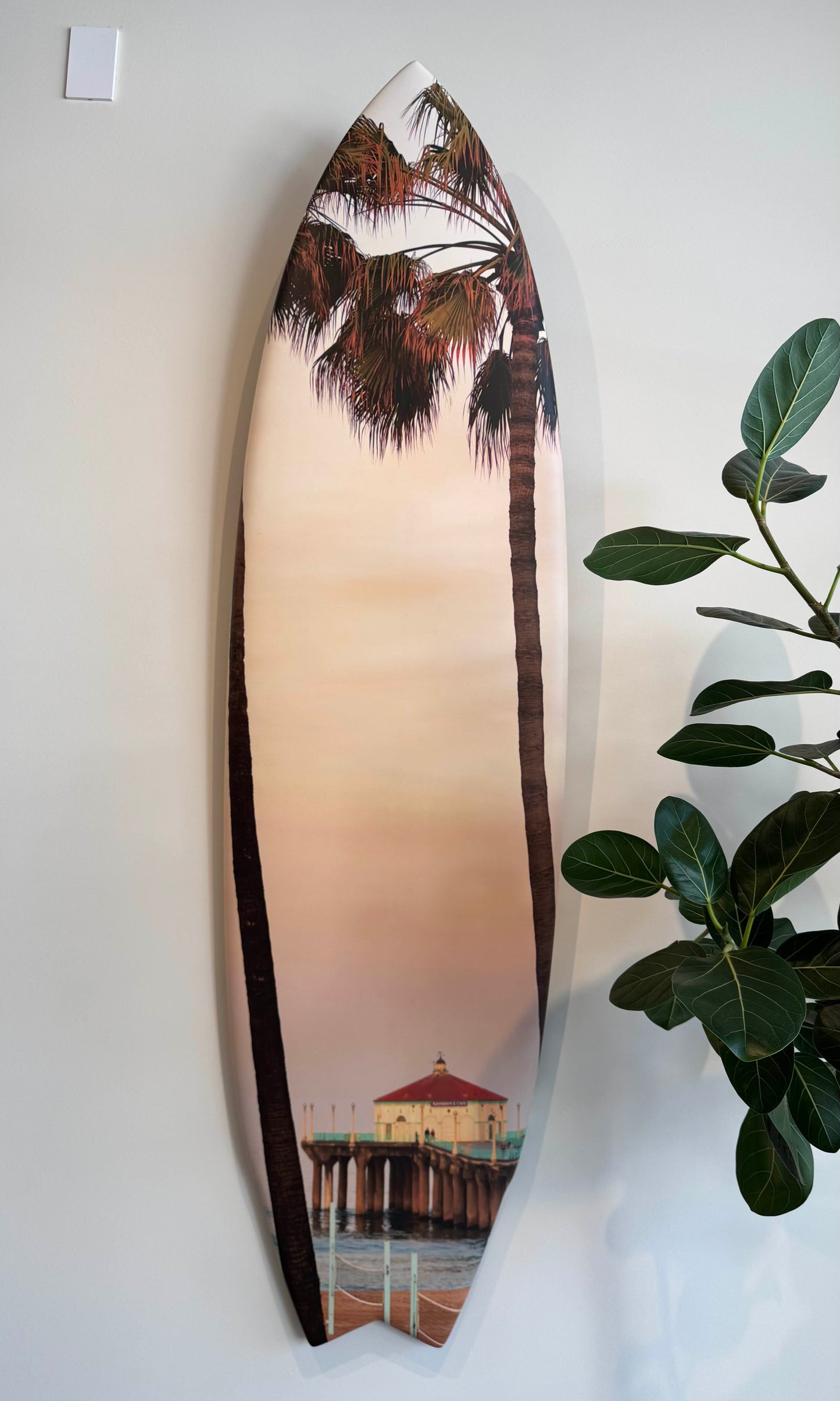 Six foot surfboard custom artwork with a photo of a palm tree and Manhattan Beach pier on a white wall.