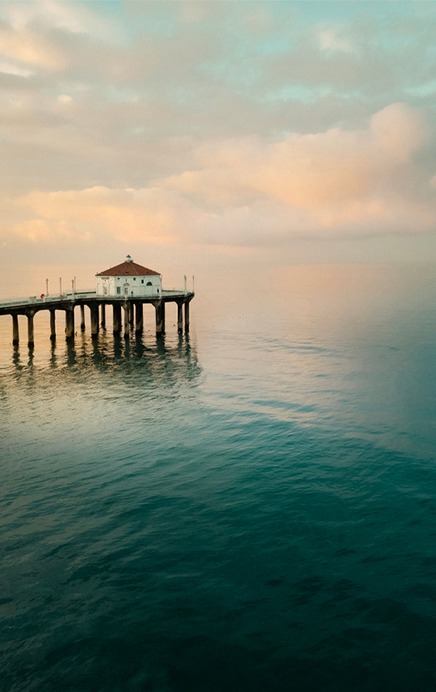 Manhattan Beach Pier with the red roof roundhouse building at the end extending into calm water under a cloudy sky.